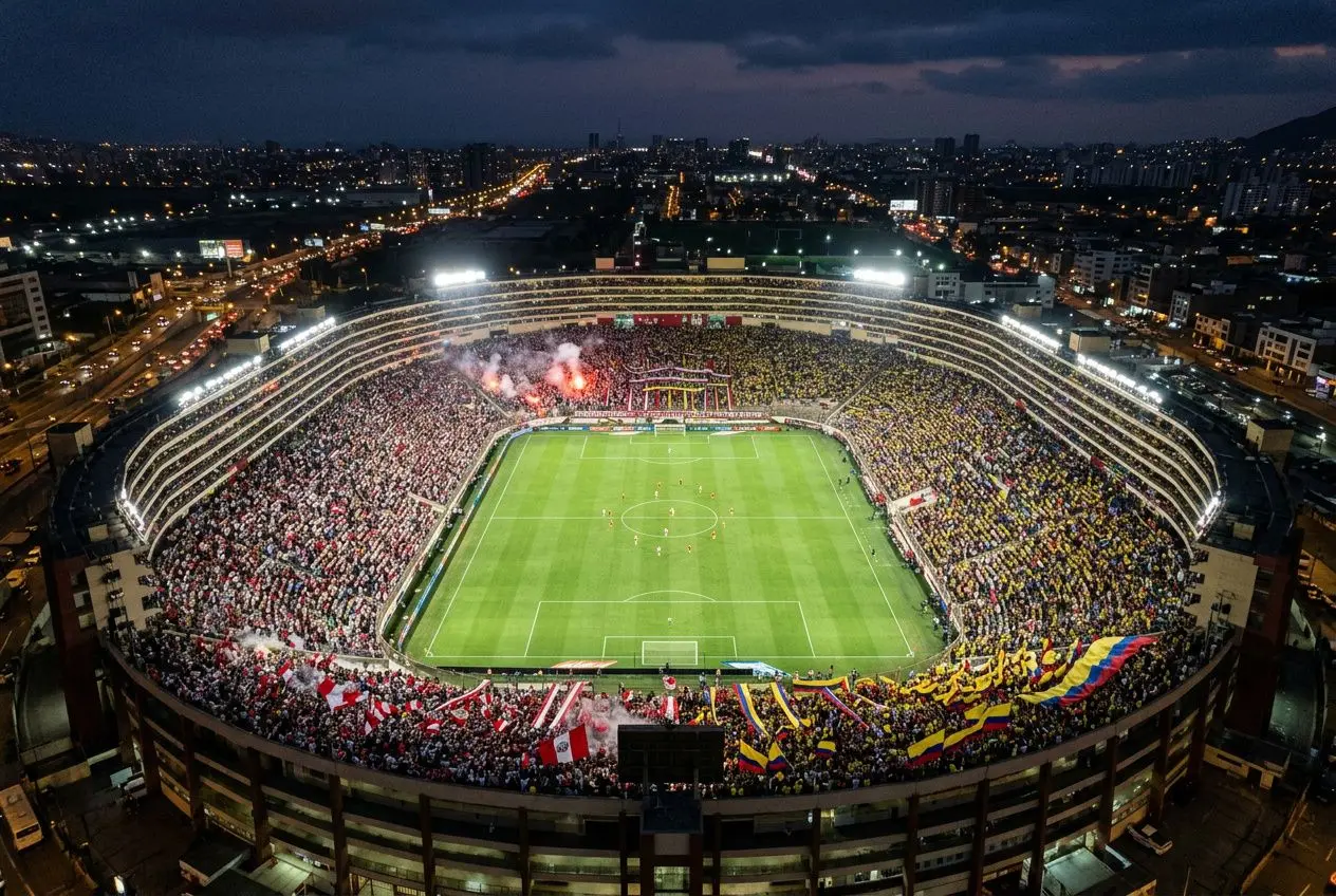 Stade plein lors d’un match de football nocturne, tifos et drapeaux colorés autour du terrain, ambiance intense en Amérique du Sud. éliminatoire coupe du monde 2026 zone amérique du sud