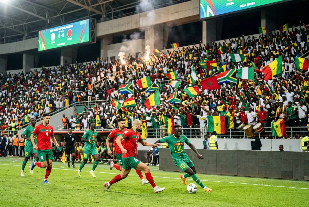 Supporters africains en tribunes agitant des drapeaux lors d’un match, joueurs en action sur le terrain. qualification coupe du monde 2026 zone afrique et ambiance festive.