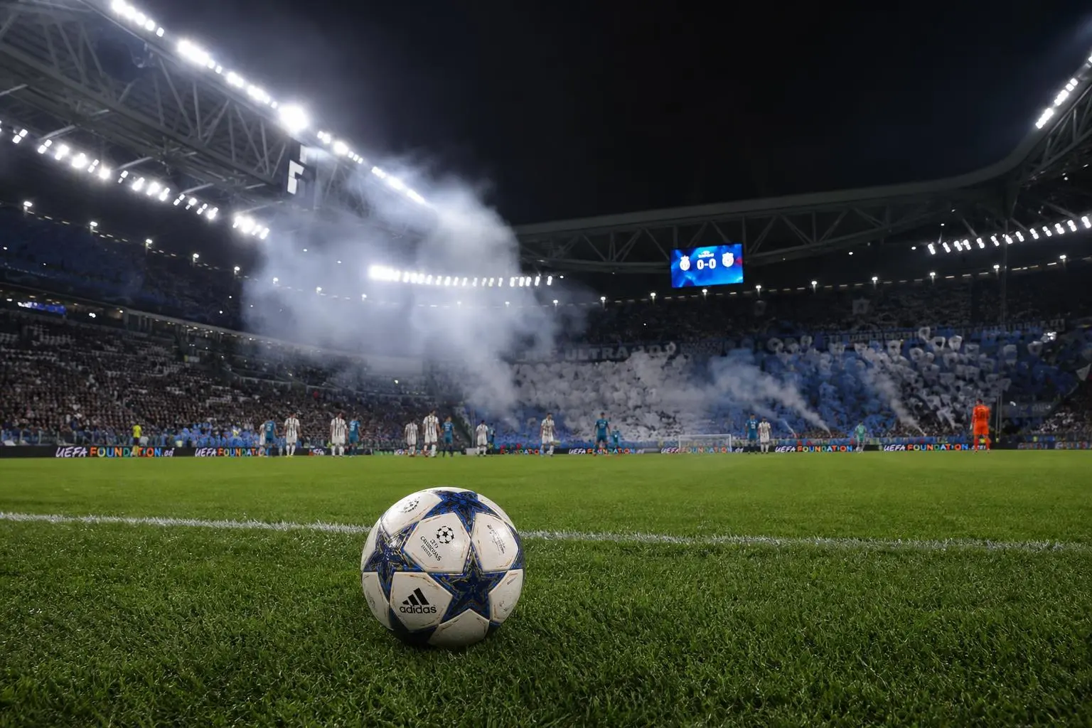 Balle de football Adidas sur le pré vert d’un stade illuminé, fumée et tribunes remplies en arrière-plan, ambiance de match de UEFA Champions League, prediction foot champions league intégrable.