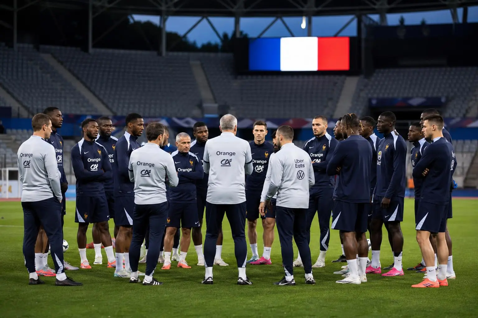 Groupe de joueurs de l'équipe de France en séance d’entraînement sur le terrain, visages concentrés, entraîneur au centre, drapeau tricolore en arrière-plan. match préparation coupe du monde 2026 calendrier france