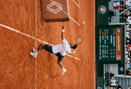 Joueur de tennis sur terre battue effectuant un smash près du filet lors d’un match, tableau des statistiques et spectateurs en arrière-plan. intégrer: comment gagner au tennis paris sportif
