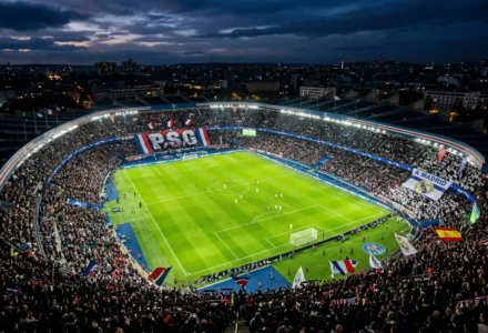 Stade rempli pour un match de football, vue aérienne du terrain vert sous les lumières, tribunes bondées avec drapeaux et banderoles, ambiance nocturne. match aller retour ligue des champions.